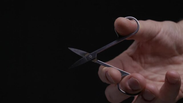 A man hand holds a small pair of metal nail scissors and closes them against a dark background. Concept of grooming, meticulous care, precision, or preparing for a task.