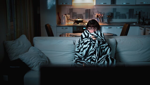 woman relaxes in the evening at home in front of the TV, brunette woman wrapped in a blanket and watches TV, switches channels, have fun and enjoy