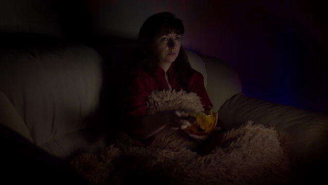 sad brunette woman sits in sofa with chips at night and watches tv with emotion, have fun and enjoy
