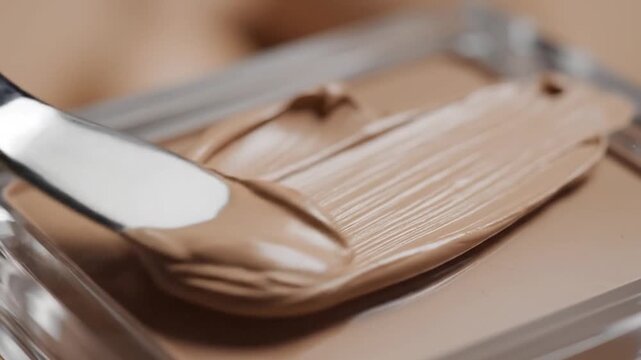 A l spatula smoothly spreads creamy beige foundation on a clear palette
