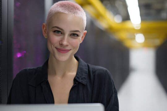 Female caucasian young pinkhair server-room technician smiling with laptop in data center aisle short buzzcut confident it specialist