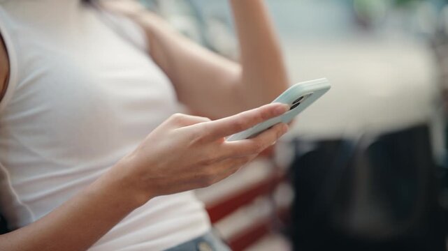 Slow motion, close up shot of a young asian woman's hands scrolling and typing on a smartphone screen while sitting on a public bench, representing connection and modern digital lifestyle