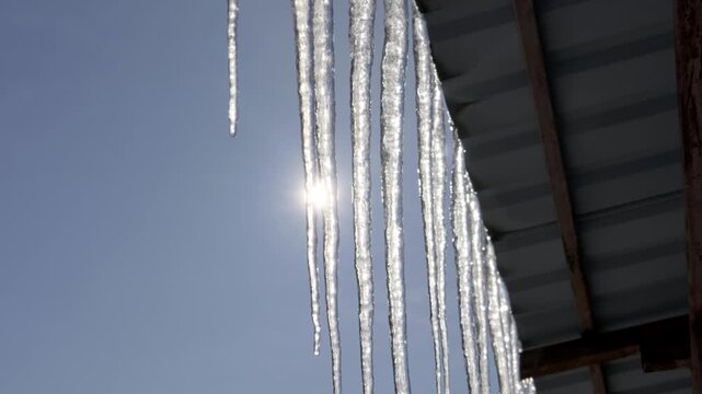 Transparent icicles hang from a metal roof edge against a blue sky, illuminated by bright sunlight with lens flare. This illustrates the concept of early spring thaw and seasonal melting ice