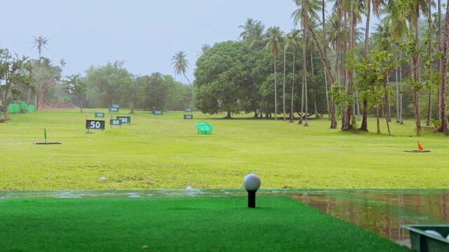 Low Angle View Of Golf Driver Striking Ball On Rainy Tropical Driving Range, Short Beginner Shot During Practice Under Wet Weather And Dark Storm Clouds