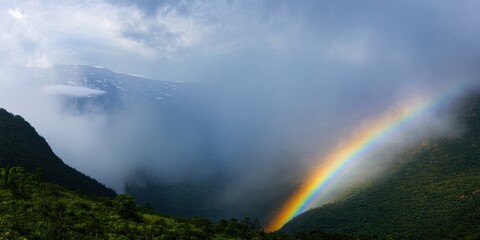 Fototapeta premium Dramatic landscape features lush hills, vibrant rainbow, misty clouds. Dark sky contrasts bright colors, creating serene view.