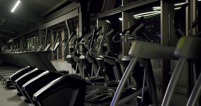 Dark interior of an empty fitness club at night with rows of modern treadmills and elliptical cross trainers standing ready for a workout, reflecting the city lights from the large windows