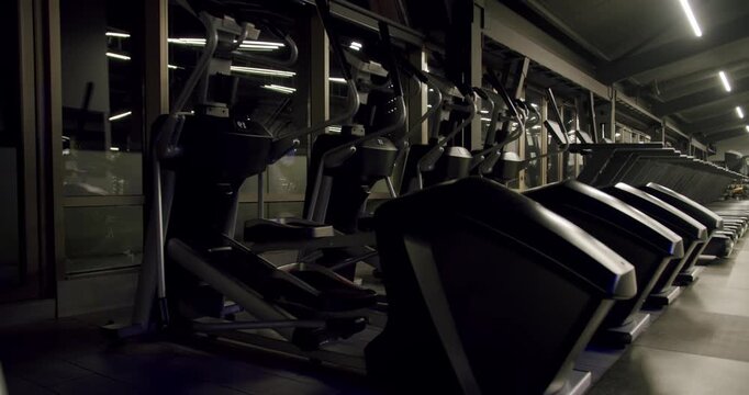 Dark and atmospheric modern gym with rows of treadmills and elliptical trainers standing ready for a workout. Fitness center interior with professional cardio equipment and moody lighting