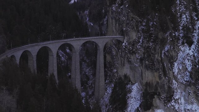 Aerial view shows Landwasser Viaduct in the Swiss Alps near Filisur as the Glacier Express crosses. Cool winter light reveals arches, conifers, rocky cliffs, and snow.
