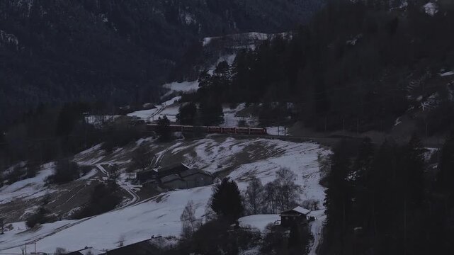Aerial view shows the red Glacier Express crossing Landwasser Viaduct in Graubunden, Switzerland, during winter, with a slow pan over snowy valley and dark forests.