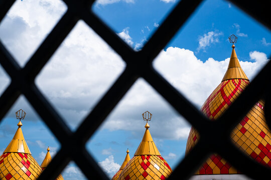 Roudhotul Muchlisin Mosque seen through a lattice against clear blue sky and cloudy with large  golden and red patterned domes during Ramadan in Jember, Indonesia.