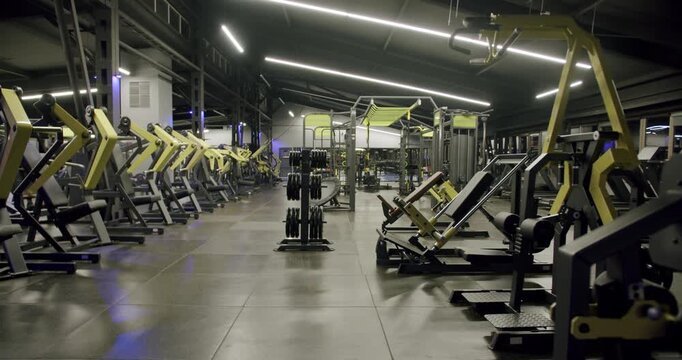 Spacious and modern fitness center interior with rows of professional workout machines and weightlifting equipment, illuminated by stylish ceiling lights in a dark, atmospheric setting