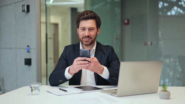 Smiling business man uses smartphone in contemporary office, displaying happiness and satisfaction. Engaged with mobile technology, finding joy and pleasure in digital interaction at workplace.