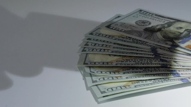 Fanned hundred dollar bills under shadow on white desk, closeup of crisp banknotes with Benjamin Franklin portrait and blue security strip, subtle creeping shadow evokes audit