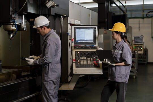 Asian adult man technician checks machine chamber with handheld controller while asian adult woman operator studies CNC panel, supporting digital workflow, real time monitoring and precision setup.