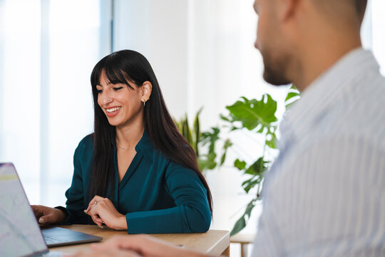 A smiling woman works on a laptop while a man sits opposite her in a bright office setting.