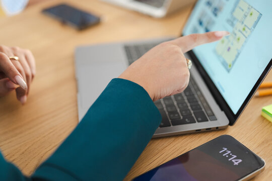 A person's hand points to a floor plan on a laptop screen, with a smartphone displaying the time on the desk.