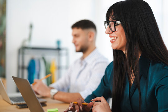 A woman wearing glasses smiles while working on a laptop, with a man in the background also engaged in work.