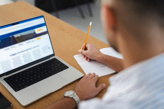 A person is taking notes in a notebook while looking at a laptop screen displaying travel information.