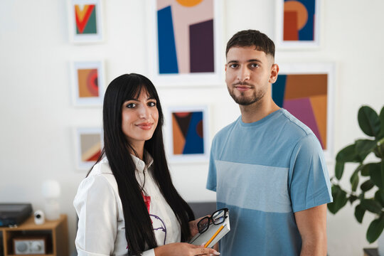 A woman and a man stand together in a modern room with abstract art on the wall. The woman holds a notebook, pencil, and glasses.