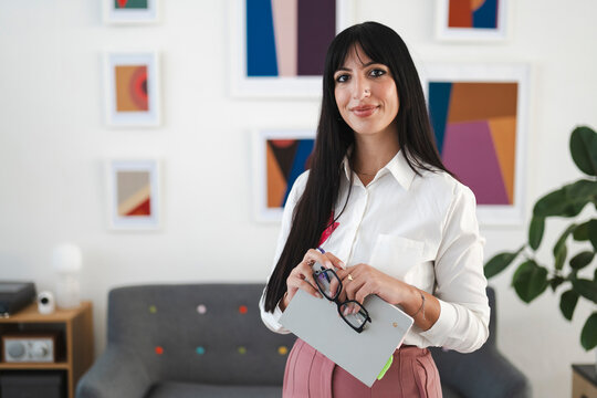 A professional woman in a white shirt and pink skirt holds a notebook and glasses, standing in a modern office with abstract art on the wall.
