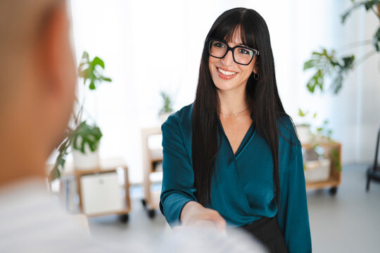 A smiling woman in glasses shakes hands with a person in the foreground, signifying a successful business meeting or introduction.
