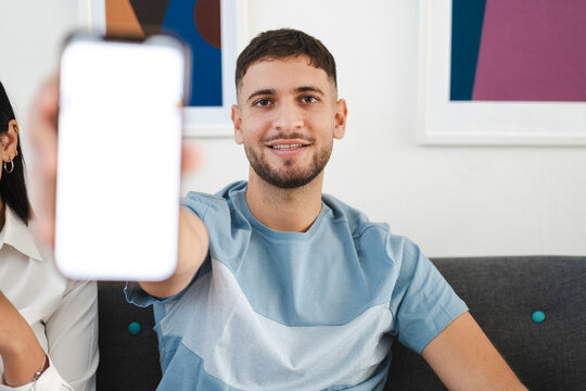 A young man smiles as he holds up a smartphone with a blank screen, inviting viewers to imagine its content.