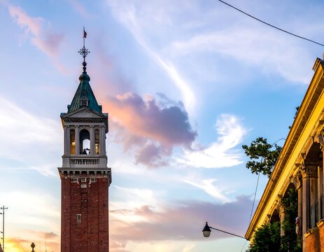 A tall, brick bell tower with a copper roof and cross stands against a pastel-colored sky, near classical architecture