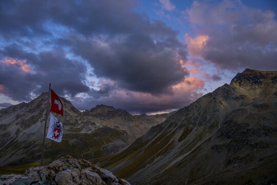 Swiss flag and flag of the SAC with cloudy sky at blue hour and Engadine mountains, St Moritz, Engadine, Grisons, Switzerland