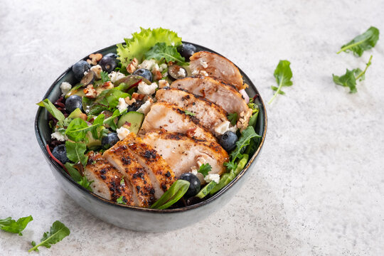 Grilled chicken and blueberry salad with greens and feta cheese arranged in a black bowl on light gray surface