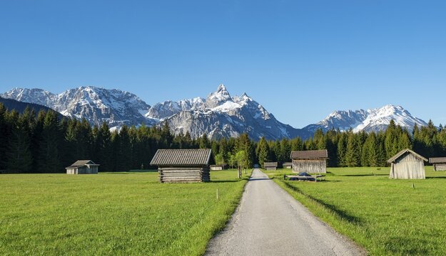 Road through meadow with hay barns, in the background snow-covered mountain peaks in spring, Mieminger Kette with Ehrwalder Sonnenspitze, Ehrwald, Tyrol, Austria