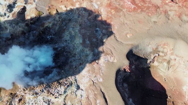 Aerial drone view of steaming geothermal vents and bubbling mud pools in a volcanic geothermal field in Iceland. Mineral rich ground in shades of beige, red, and blue surrounds active fumaroles releas