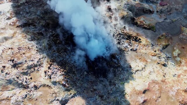Aerial drone view of steaming geothermal vents and bubbling mud pools in a volcanic geothermal field in Iceland. Mineral rich ground in shades of beige, red, and blue surrounds active fumaroles releas