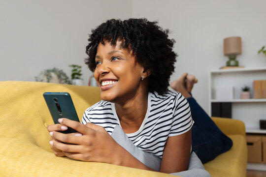 Smiling young black woman using mobile phone relaxing on sofa at home. Technology and gen z people concept