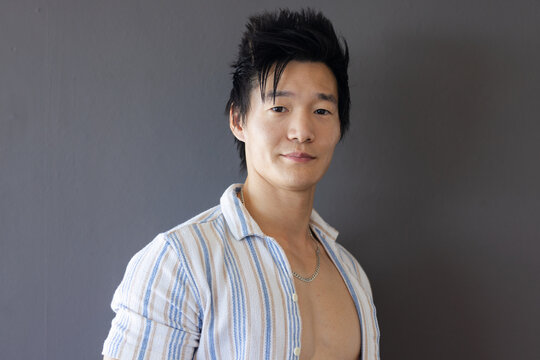 Asian man posing against plain gray wall in studio, wearing open striped shirt and silver chain