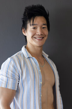Asian man posing and smiling, wearing open striped shirt with silver chain against dark gray wall