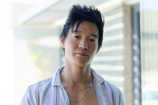 Vertical video: Facing Asian man wearing striped shirt and silver chain on balcony with louvers