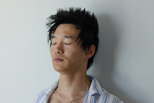 Adult Asian man leaning against pale wall in studio closing eyes wearing striped shirt silver chain