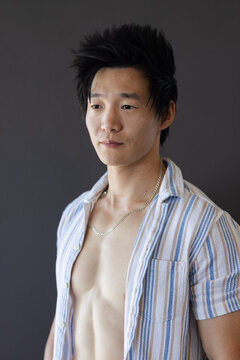 Asian man posing in studio with averted gaze, showing open striped shirt and silver chain