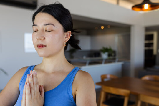 Asian woman praying, standing in apartment kitchen by wooden table wearing blue tank top