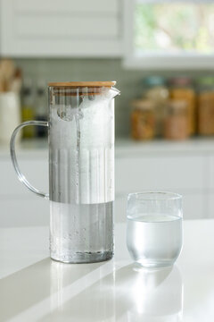 Showing glass pitcher with wooden lid condensing and foaming on glossy kitchen countertop, tumbler