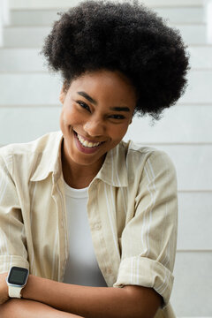 Woman sitting on pale steps leaning, smiling, wearing beige shirt, white tank and smartwatch