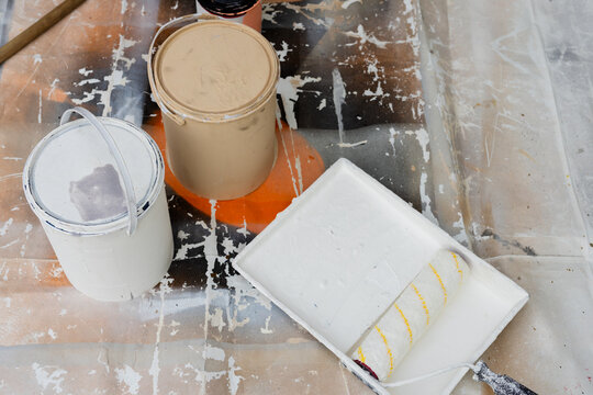 Displaying paint cans and tray sitting on protective plastic sheet at garage, with roller and stick