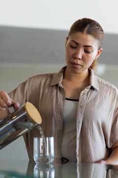 Pouring woman in beige shirt and ribbed top at kitchen counter, steadying, with pitcher and glass