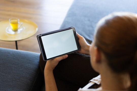 Female-presenting adult sitting on sofa holding tablet in case, dark cushion, glass cup, copy space