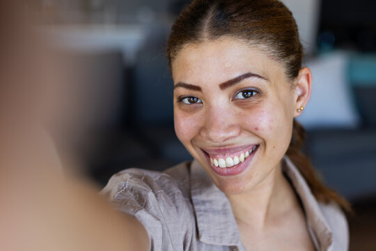 Smiling African woman taking selfie in living room, holding smartphone with beige shirt, gold studs