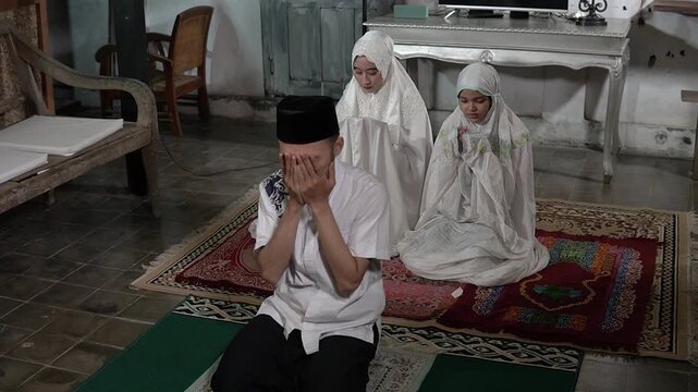 Asian father supplication leading family prayer, man in cap with hands raised while two girls in white hijab sit behind, humble