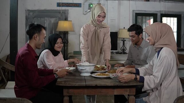 Asian muslim friends sharing warm iftar meal around wooden table, hijab host placing platter of rice and dishes, cozy lamp light,