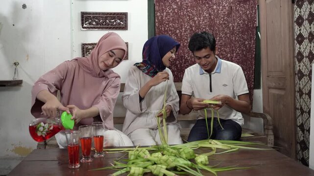 Asian trio preparing festive table, three friends in hijab and casual shirt weaving palm pouches, table full of folded pouches
