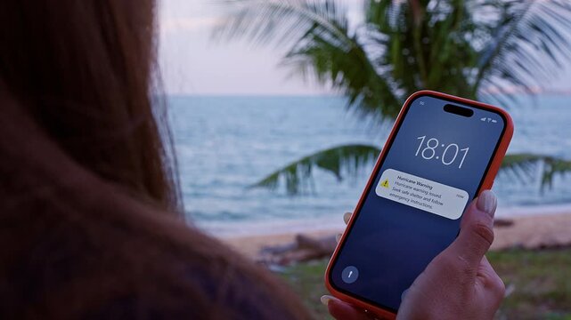 Young Woman Standing on Tropical Beach Holding Smartphone Showing Hurricane Warning Alert Notification While Looking Toward Ocean During Dangerous Storm Situation