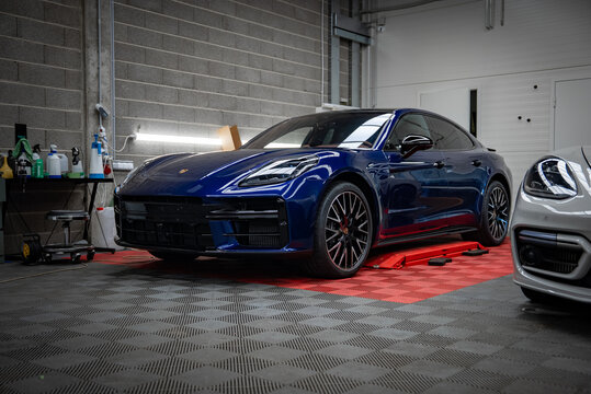 Deep blue Porsche Panamera 4 sits on a red hydraulic platform in an indoor workshop with gray cinderblock walls, bright tube lighting, and shelves of tools and bottles.
